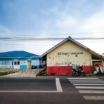 Two low school buildings along a street with a zebra crossing in the foreground; the cream building has graffiti and reads 'SD Negeri Cendrawas...' with a motorcycle nearby.