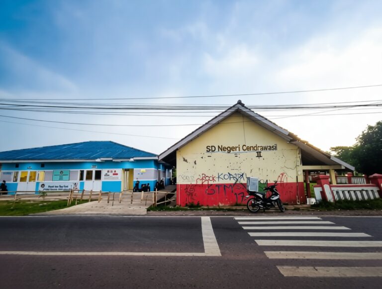 Two low school buildings along a street with a zebra crossing in the foreground; the cream building has graffiti and reads 'SD Negeri Cendrawas...' with a motorcycle nearby.