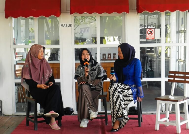 Three women in hijabs seated on a small stage outdoors; one speaks into a microphone while the others listen, under a red canopy.