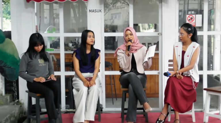 Panel of four women seated indoors; the second woman from left listens as the hijab-wearing speaker raises a microphone.