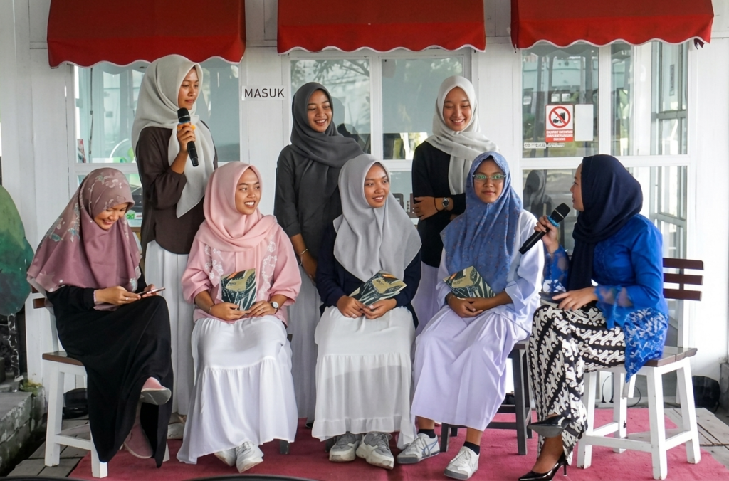 A group of seven women wearing hijabs sits and chats on a pink rug, one woman speaks into a microphone while others hold programs.