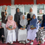 A group of seven women wearing hijabs sits and chats on a pink rug, one woman speaks into a microphone while others hold programs.