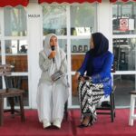 Two women in hijabs sit on a red carpeted stage, one holding a microphone as they chat.