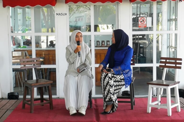 Two women in hijabs sit on a red carpeted stage, one holding a microphone as they chat.