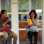 Two women seated on stools, each holding a microphone during a discussion; one reads from a yellow booklet.