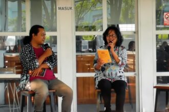 Two women seated on stools, each holding a microphone during a discussion; one reads from a yellow booklet.