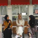 Three women in hijabs sit on stools outdoors, each holding a microphone as they discuss on a panel.