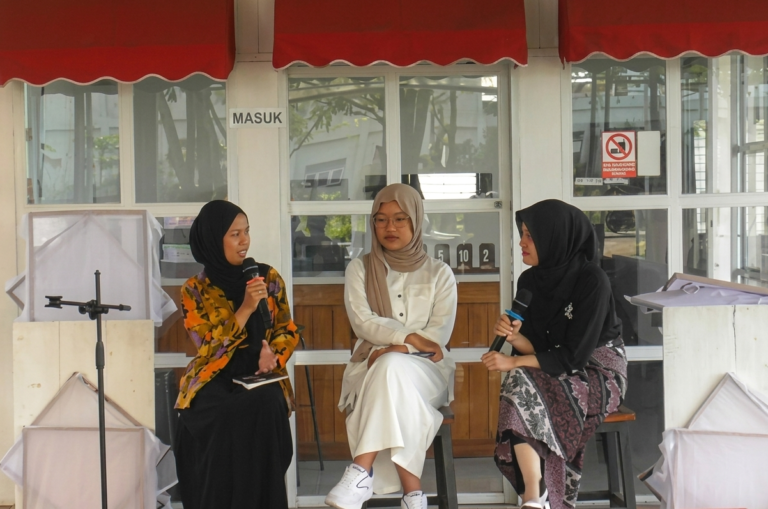 Three women in hijabs sit on stools outdoors, each holding a microphone as they discuss on a panel.
