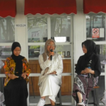 Three women wearing hijabs sit on a stage; the middle woman speaks into a microphone while the others listen or hold notes.