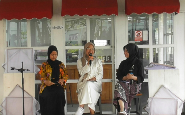 Three women wearing hijabs sit on a stage; the middle woman speaks into a microphone while the others listen or hold notes.