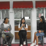 Panel discussion with three people seated on stage in front of glass doors and red awnings; center woman speaks into a microphone while others listen.