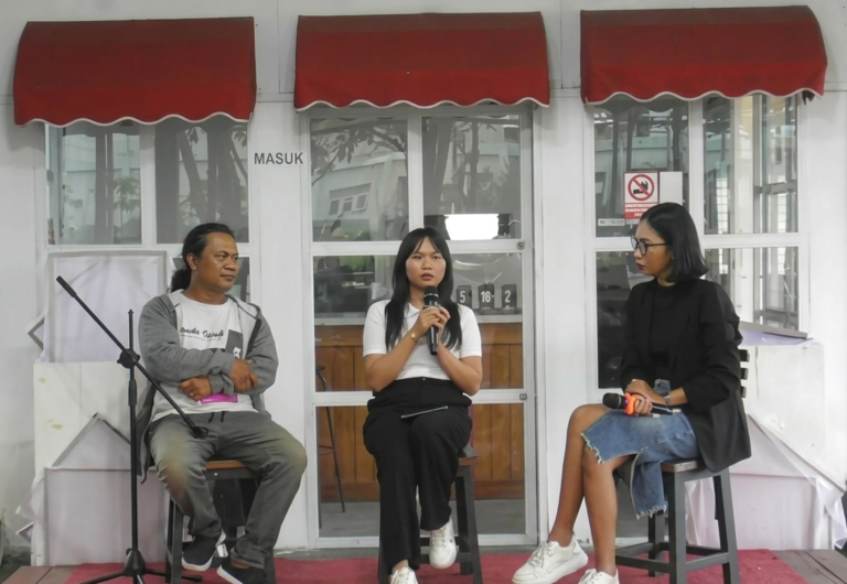 Panel discussion with three people seated on stage in front of glass doors and red awnings; center woman speaks into a microphone while others listen.