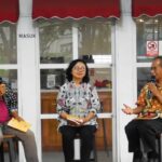 Three panelists sit on stools in front of a glass storefront, each holding a microphone during an outdoor discussion.