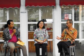 Three panelists sit on stools in front of a glass storefront, each holding a microphone during an outdoor discussion.