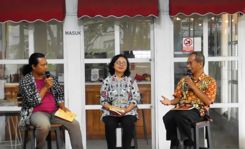 Three panelists sit on stools in front of a glass storefront, each holding a microphone during an outdoor discussion.