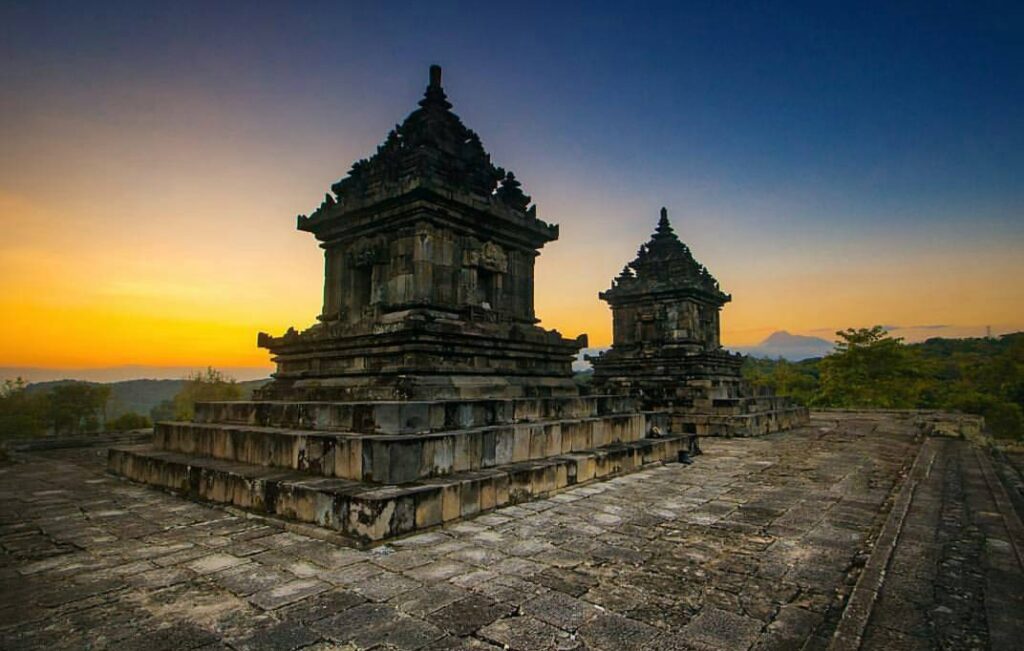 Senja di Candi Barong YogyakartaSalah satu referensi tempat yang perlu kamu kunjungi ketika lib - Mabur.co Two ancient stone temple towers on a raised, stepped platform at sunset with a warm orange sky behind them.