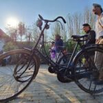 A vintage bicycle in the foreground as a group of people gathers in a sunny courtyard behind it.
