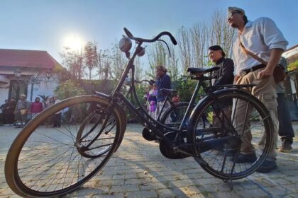 A vintage bicycle in the foreground as a group of people gathers in a sunny courtyard behind it.