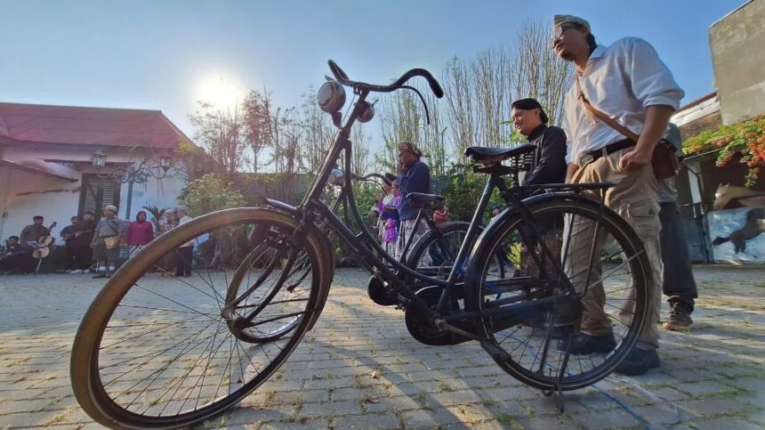A vintage bicycle in the foreground as a group of people gathers in a sunny courtyard behind it.