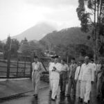 Group of men in light-colored suits and traditional caps walking along a wet road beside a wooden fence, with hills/volcano in the background.