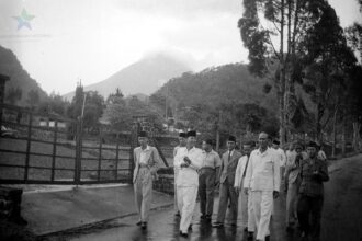 Group of men in light-colored suits and traditional caps walking along a wet road beside a wooden fence, with hills/volcano in the background.