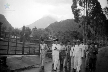 Group of men in light-colored suits and traditional caps walking along a wet road beside a wooden fence, with hills/volcano in the background.