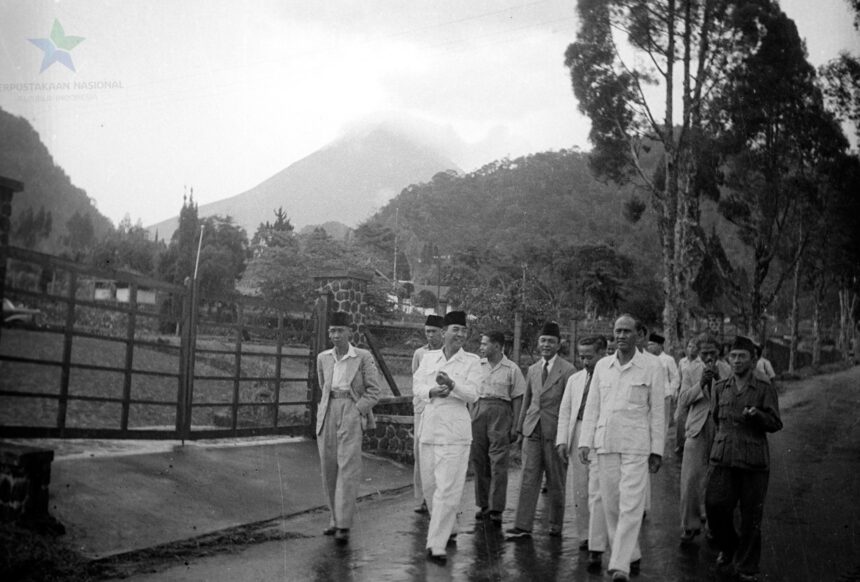 Group of men in light-colored suits and traditional caps walking along a wet road beside a wooden fence, with hills/volcano in the background.