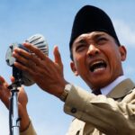 Man in a tan suit and black hat speaks into a vintage microphone outdoors, gesturing with his hands.
