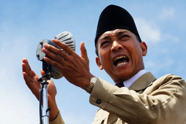 Man in a tan suit and black hat speaks into a vintage microphone outdoors, gesturing with his hands.