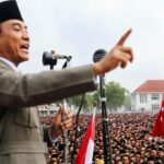 Politician in a suit and black cap addressing a massive crowd at a rally, with microphones and red-and-white flags nearby.