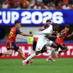 Soccer players chase the ball in a crowded stadium, a player in red and gold pulls on another player's white kit.