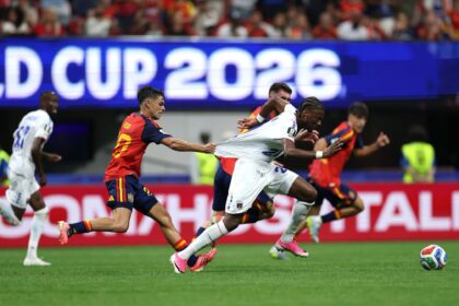 Soccer players chase the ball in a crowded stadium, a player in red and gold pulls on another player's white kit.