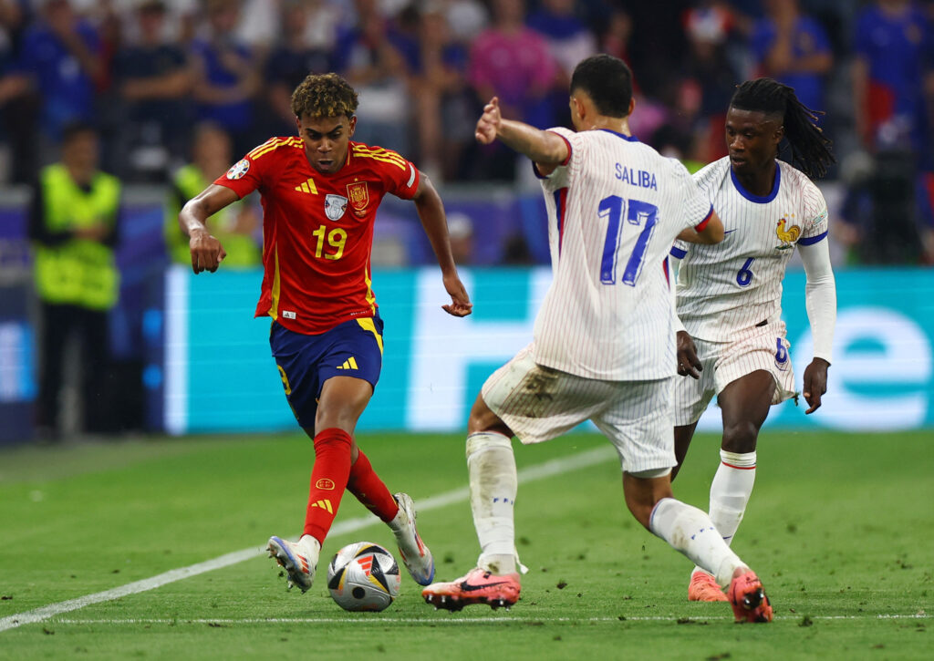 Spanish player (number 19) in red kit dribbles the ball as French defenders in white striped kits contest him on the field with a cheering crowd in the background.