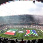 Bird's-eye view of a full stadium with a circular field display featuring large national flags and cheering spectators.