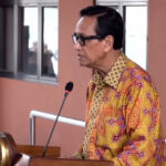 Man in a patterned shirt speaks at a podium with a microphone in a conference setting.