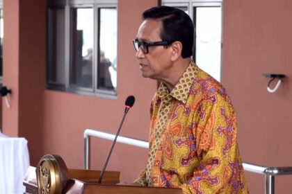 Man in a patterned shirt speaks at a podium with a microphone in a conference setting.