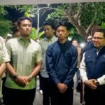 Group of men in smart casual and jackets posing for a photo in a hallway with greenery in the background, forming a formal line up.
