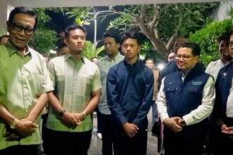 Group of men in smart casual and jackets posing for a photo in a hallway with greenery in the background, forming a formal line up.