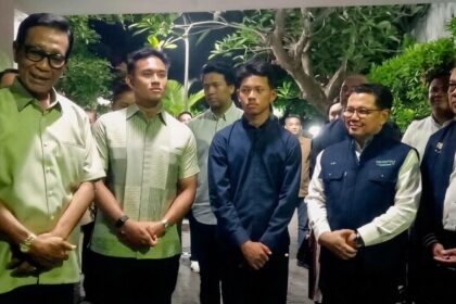 Group of men in smart casual and jackets posing for a photo in a hallway with greenery in the background, forming a formal line up.