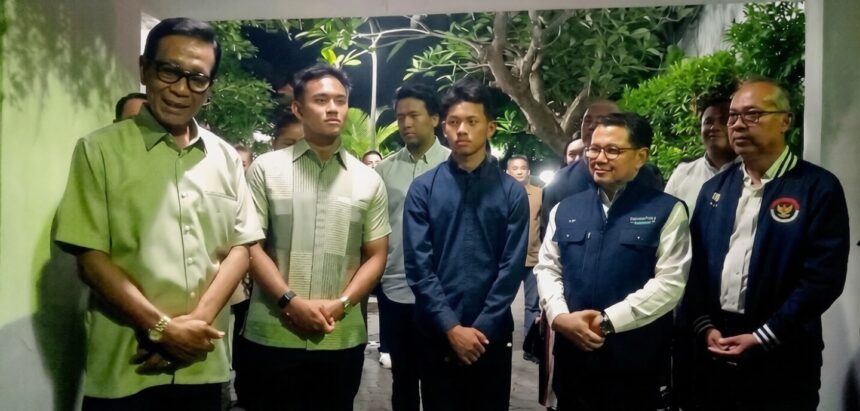 Group of men in smart casual and jackets posing for a photo in a hallway with greenery in the background, forming a formal line up.