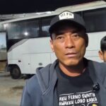 Man wearing a black cap and gray jacket speaks to the camera in front of a white bus at night, with a boy in a blue jacket behind him.