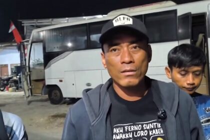 Man wearing a black cap and gray jacket speaks to the camera in front of a white bus at night, with a boy in a blue jacket behind him.