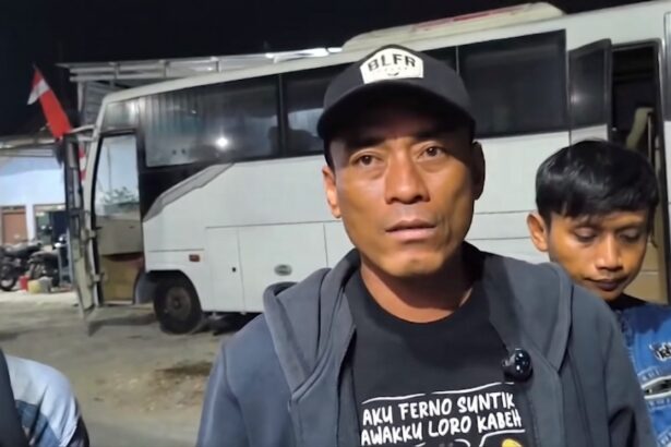 Man wearing a black cap and gray jacket speaks to the camera in front of a white bus at night, with a boy in a blue jacket behind him.