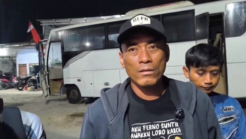 Man wearing a black cap and gray jacket speaks to the camera in front of a white bus at night, with a boy in a blue jacket behind him.