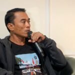 Man speaking into a handheld microphone while seated in a chair, wearing a dark hoodie and black T-shirt.