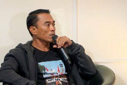 Man speaking into a handheld microphone while seated in a chair, wearing a dark hoodie and black T-shirt.