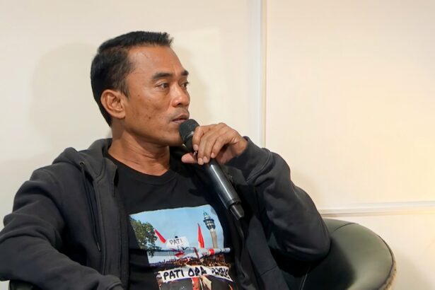 Man speaking into a handheld microphone while seated in a chair, wearing a dark hoodie and black T-shirt.