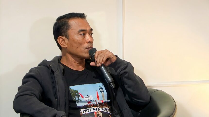 Man speaking into a handheld microphone while seated in a chair, wearing a dark hoodie and black T-shirt.