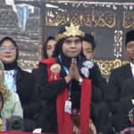 Person in black attire with a golden crown and red scarf stands with hands pressed together in a prayer pose on a stage, surrounded by formally dressed attendees.