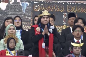 Person in black attire with a golden crown and red scarf stands with hands pressed together in a prayer pose on a stage, surrounded by formally dressed attendees.